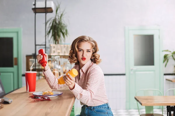 Young lady in shirt sitting at the bar counter with ketchup and mustard bottles in hands and happily looking in camera while spending time in cafe