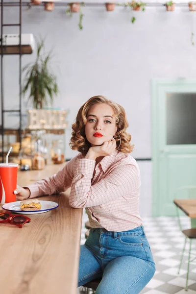 Young thoughtful lady in shirt and jeans sitting at the bar counter with coca cola glass and dremily looking in camera in cafe