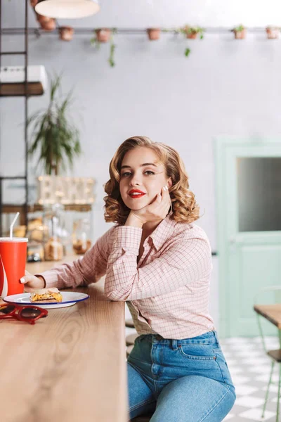 Beautiful lady in shirt and jeans sitting at the bar counter with coca cola glass and dremily looking in camera in cafe