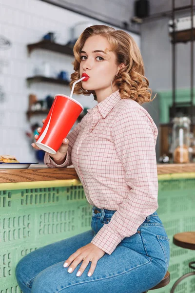 Young beautiful lady in shirt and jeans sitting at the bar counter and drinking soda water while thoughtfully looking in camera in cafe