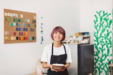Siyah önlük ve beyaz T-shirt el yapımı tabak mutlu kamera seyir Çanak Atölyesi elinde tutan sevimli gülümseyen kız