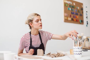 Siyah önlük ve çizgili tişört düşünceli Çanak Atölyesi araçları seçme kil ile masada oturan güzel kız