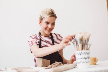 Gülümseyen kız siyah önlük ve mutlu bir şekilde modern Çanak Atölyesi araçları seçme kil ile oturan çizgili tişört