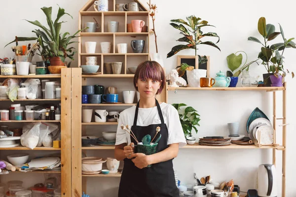 Beautiful girl with colorful hair in black apron and white T-shirt holding handmade bowl with plant in hands dreamily looking in camera at pottery studio