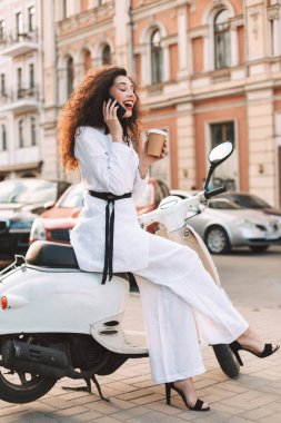 Beyaz kostüm fincan kahve ve sevinçle arka plan üzerinde şehir manzaralı onu telefonda konuşurken ile beyaz motosiklet üzerinde oturan siyah kıvırcık saçlı Genç Bayan