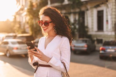 Gülümseyen güzel Bayan güneş gözlüğü ve mutlu bir şekilde sokakta duran cep telefonunu kullanarak beyaz ceket siyah kıvırcık saçlı