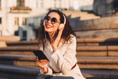 Genç gülümseyen Bayan güneş gözlüğü ve beyaz ceket elinde cep telefonu ile merdivenlerde oturan siyah kıvırcık saçlı ve kulaklık sokakta sersem dinleme müzik ile 