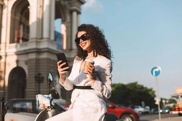 Mutlu gülümseyen kadın güneş gözlüğü ve beyaz kostüm arka plan üzerinde şehir manzaralı fincan kahve ve elinde cep telefonu ile beyaz motosiklet üzerinde oturan siyah kıvırcık saçlı