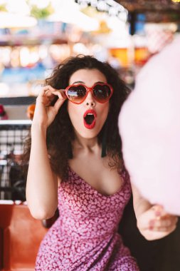 Güneş gözlüğü ve pamuk şeker holding ve amazedly eğlence parkı zaman harcama süre kamera seyir elbise siyah kıvırcık saçlı güzel Bayan