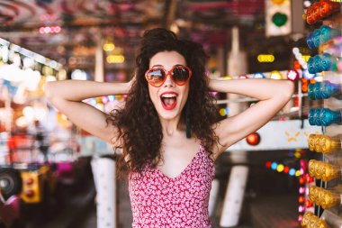 Güneş gözlüğü ve mutlu bir şekilde kamera eğlence parkı konumlar arka plan üzerinde dururken seyir elbise siyah kıvırcık saçlı genç hanımefendi