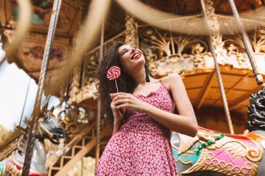 Elinde lolly pop candy ile ayakta ve mutlu bir kenara arıyorum güzel carousel ile arka plan üzerinde elbise siyah kıvırcık saçlı Genç güzel Bayan