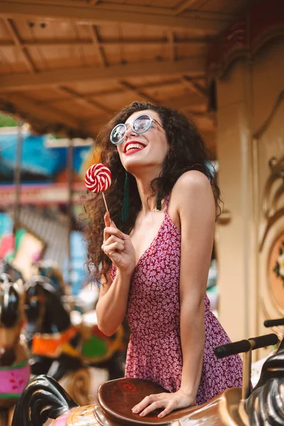 Joyful lady with dark curly hair in sunglasses and dress standing with lolly pop candy in hand and dreamily looking aside while riding on carousel in amusement park