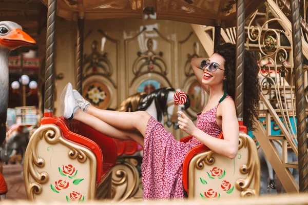 サングラスとドレス キャンディー ポップなキャンディを手に保持し ながら遊園地で楽しく過ごすカルーセルに乗って暗い巻き毛のうれしそうな女の子 — ストック写真