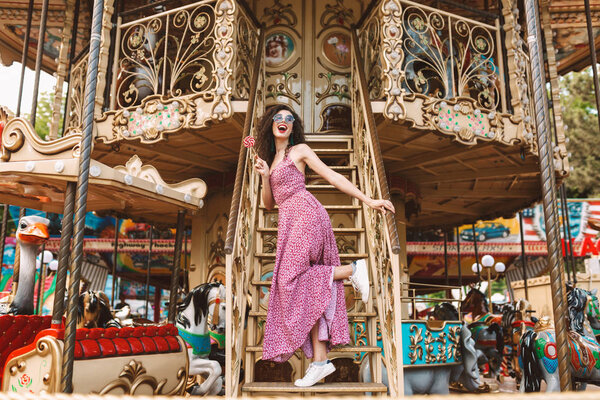 Beautiful smiling lady with dark curly hair in sunglasses and dress standing with lolly pop candy in hand and happily looking aside while spending time on carousel 