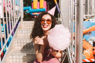 Güneş gözlüğü ve pamuk şeker el ve mutlu bir şekilde eğlence parkı zaman harcama ile oturan Doğum günü kap karanlık kıvırcık saçlı hanımefendi 