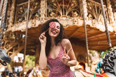 Joyful lady with dark curly hair in dress standing and covering her eye with lolly pop candy while happily looking in camera with beautiful carousel on background clipart
