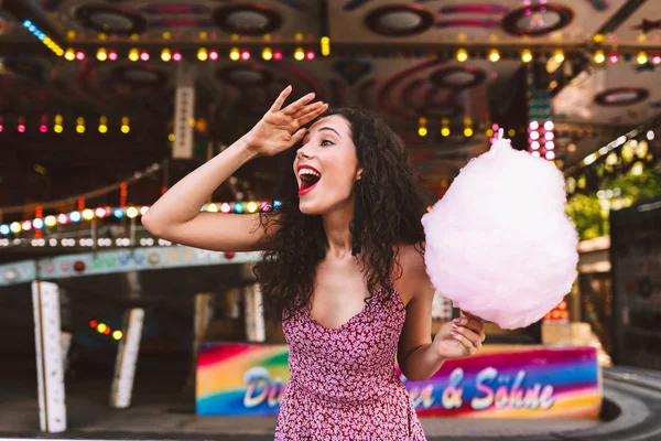 ピンクの綿飴を手にし喜んで背景にアトラクション遊園地で時間を過ごしながらよそ見をドレス状態で暗い巻き毛を持つ若いジョイフル レディー — ストック写真