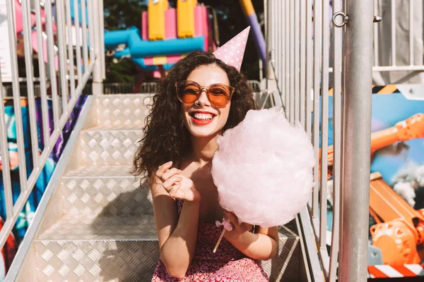 Joyful lady with dark curly hair in sunglasses and birthday cap sitting with cotton candy in hand and happily spending time in amusement park