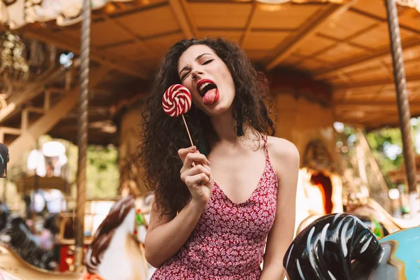 キャンディー ポップなキャンディ遊園地で背景にカルーセルで舌をカメラで見せて 手をドレス状態で暗い巻き毛を持つクールなお嬢様 — ストック写真