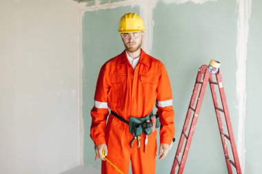 Turuncu genç Oluşturucu iş giysileri ve sarı hardhat rulet düşünceli kamera ile merdiven arka plan üzerinde arıyorum elinde tutan 