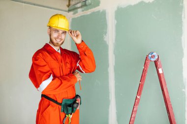Turuncu genç Foreman'ın iş giysi ve sarı hardhat sevinçle kamera ile merdiven arka plan üzerinde seyir 