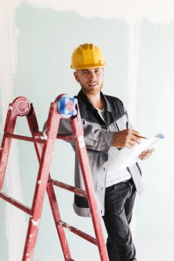 İş elbiseleri ve sarı hardhat sersem kamera tutarak plan yeni dairelerin iş yerinde kırmızı merdiven yaslanmış seyir genç mühendis