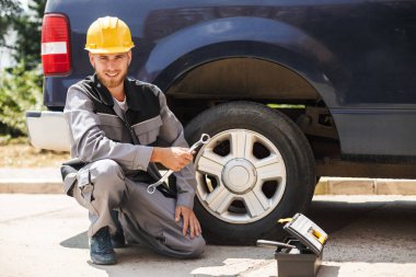 Genç gülümseyen mekanik iş elbiseleri ve sarı hardhat mutlu kamera tutarak anahtarı açık araba tekerleği değiştirmek için arıyorum