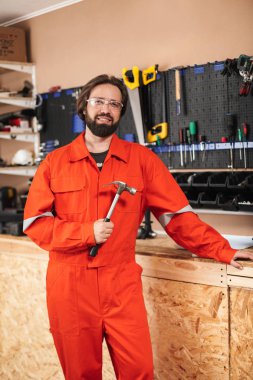 Foreman turuncu iş elbiseleri ve mutlu bir şekilde kamera araçları ile arka plan atölye üzerinde seyir çekiç tutarak koruyucu gözlük gülümseyen