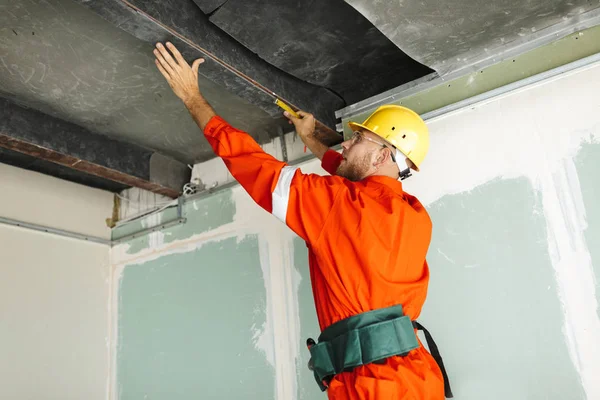 Turuncu iş elbiseleri ve sarı kask yeni daire tavanda Onarım için ölçüm bandı kullanarak genç Oluşturucu