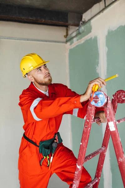 Turuncu iş elbiseleri ve düşünceli bir kenara dairede arıyorum elinde kaset ölçme ile merdiven üzerinde duran sarı hardhat genç ustabaşı