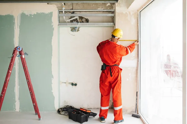 Builder in orange work clothes and yellow hardhat using measuring tape near big window in new apartment at work