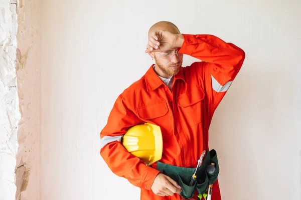 Tired foreman in orange work clothes and protective eyewear holding yellow hardhat in hand over white background