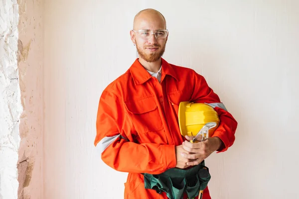 Young foreman in orange work clothes and protective eyewear holding yellow hardhat and tools in hand happily looking in camera over white backgroun