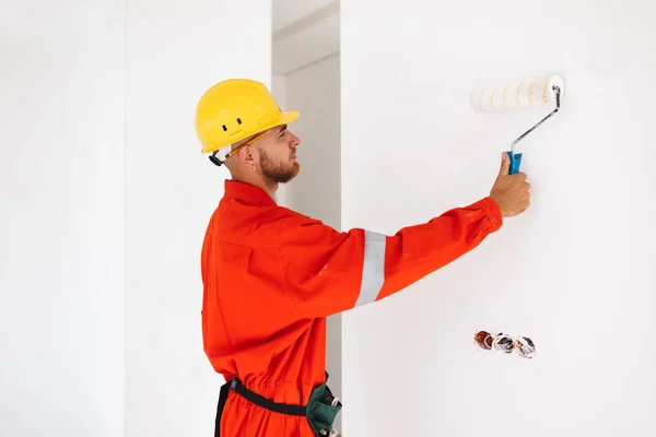 Young foreman in orange work clothes and yellow hardhat using painting roller in new apartment at work