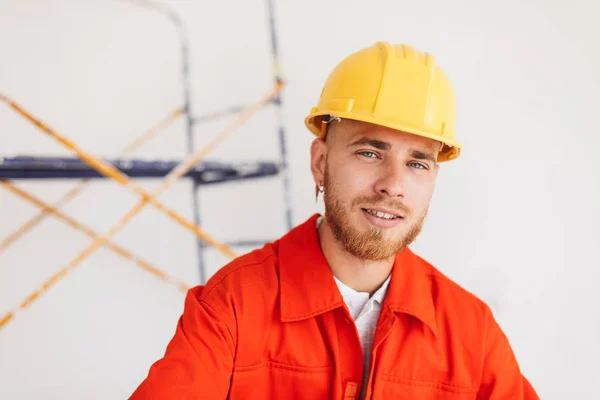 Portrait of young builder in orange work clothes and yellow hardhat with pencil behind ear dreamily looking in camera with scaffolding on background