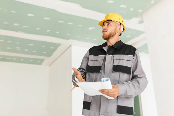Young engineer in work clothes and yellow hardhat holding plan of new apartment in hand thoughtfully looking aside at work