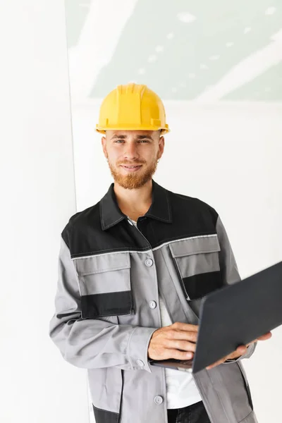 Smiling engineer in work clothes and yellow hardhat happily looking in camera holding laptop in hand over white background