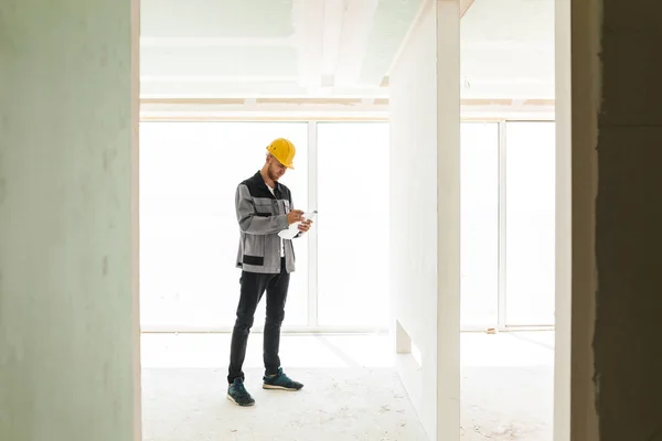 Young engineer in work clothes and yellow hardhat working with plan of new apartments with big window on background