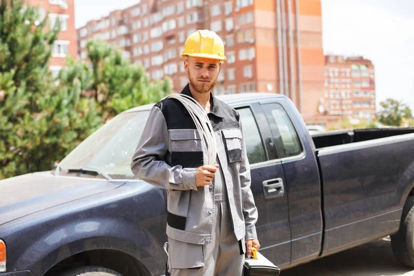 Young mechanic in work clothes and yellow hardhat holding toolbox in hand thoughtfully looking in camera with black car on background