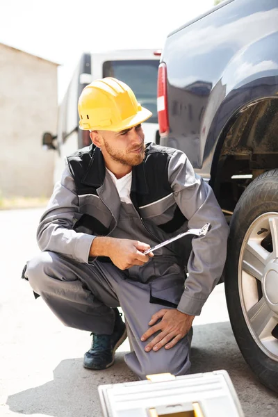 Young mechanic in work clothes and yellow hardhat thoughtfully looking aside holding wrench near car at work