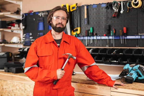 Foreman in orange work clothes and protective eyewear holding hammer dreamily looking in camera with tools on background in workshop