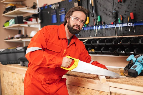 Foreman in orange work clothes and protective eyewear thoughtfully looking in camera using handsaw in workshop