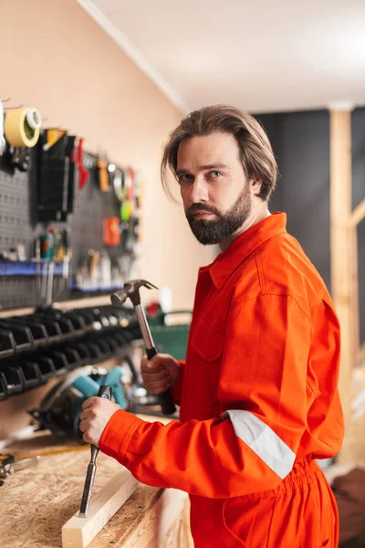 Builder in orange work clothes thoughtfully looking in camera holding hammer in workshop