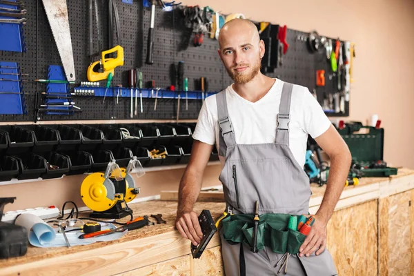 stock image Young foreman in work clothes holding stapler in hand thoughtfully looking in camera with stand of different tools on background in workshop