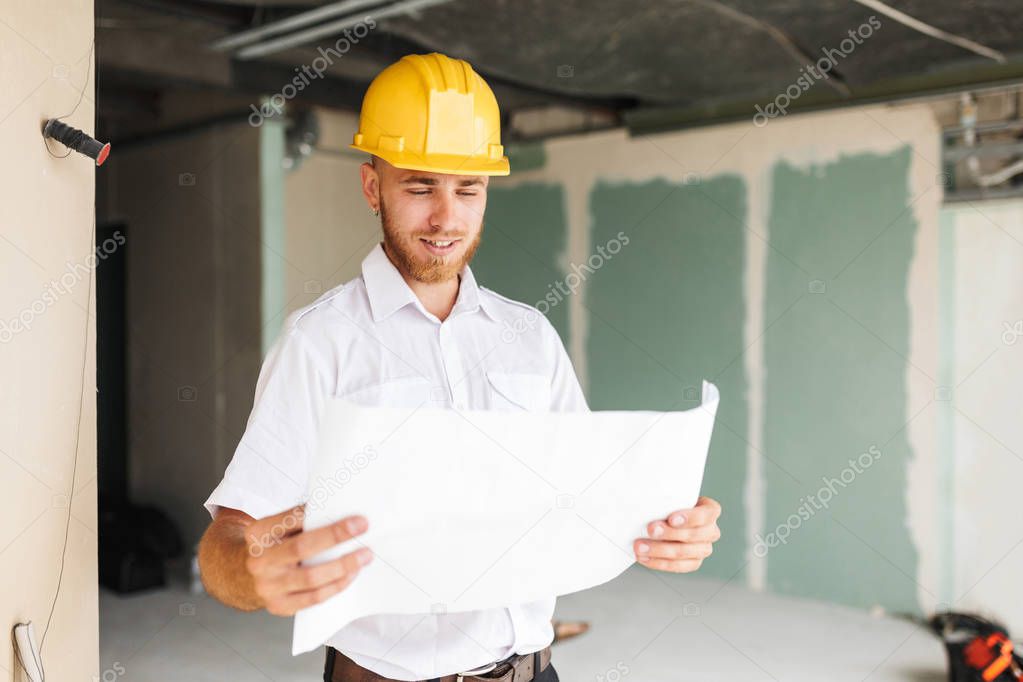 Young architect in white shirt and yellow hardhat happily looking on plan of new apartment at work