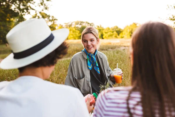 美丽的微笑的女孩拿着果汁在手愉快地谈话与朋友花时间在公园野餐 — 图库照片