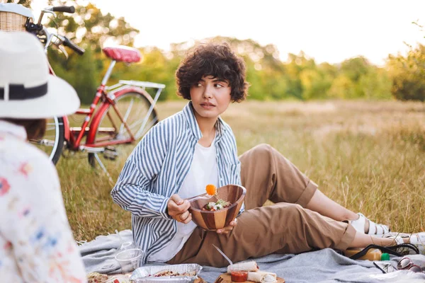 美丽的女孩在条纹衬衫拿着碗沙拉朦胧地看一边在公园野餐 — 图库照片
