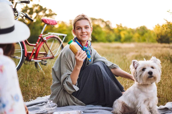 美丽的微笑女孩拿着冰壶在手愉快地看在照相机与小可爱的狗在公园野餐 — 图库照片
