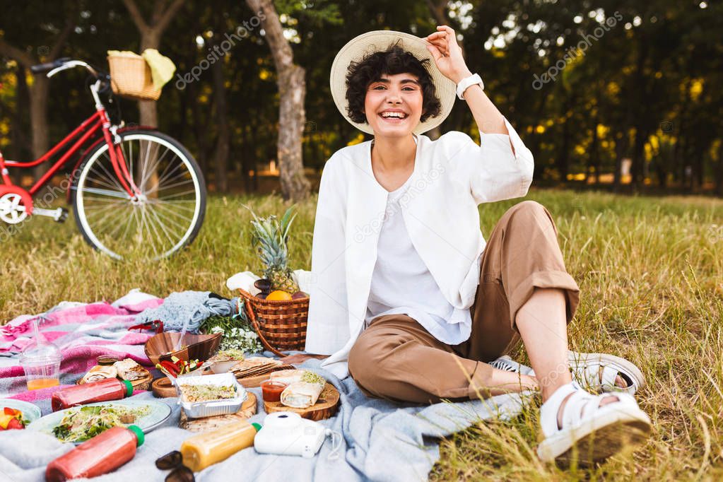 Pretty laughing girl sitting in hat and white shirt happily looking in camera spending time on picnic in park