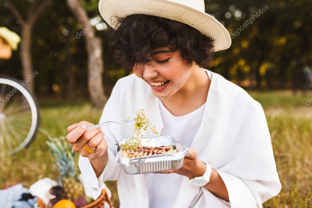 Portrait of beautiful girl in hat and white shirt eating delicious food happily spending time on picnic in park